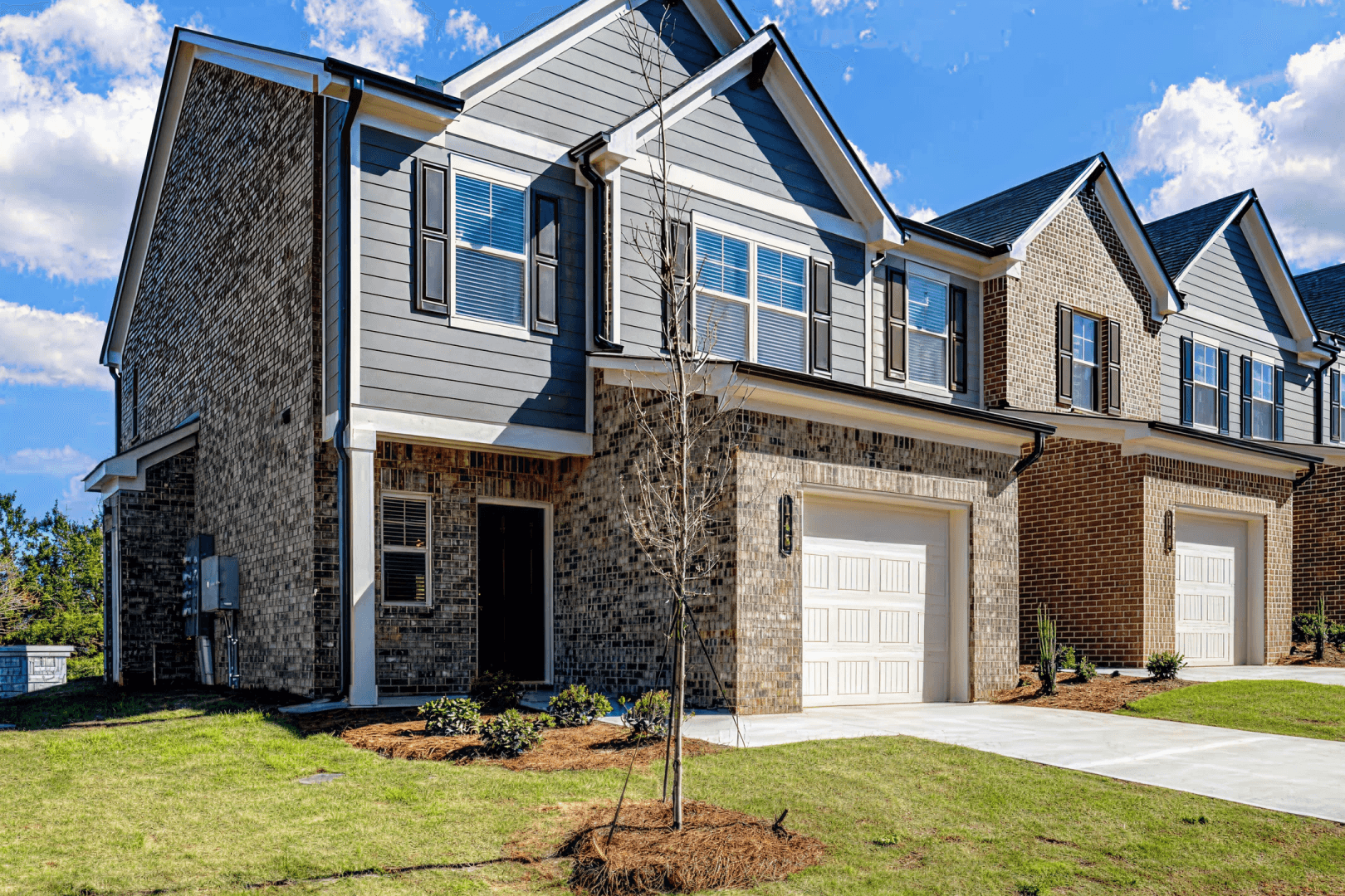 Modern Townhomes with Brick and Siding Exterior Row of newly constructed two-story townhomes featuring brick facades, gray siding, white garage doors, and young landscaping under blue sky.