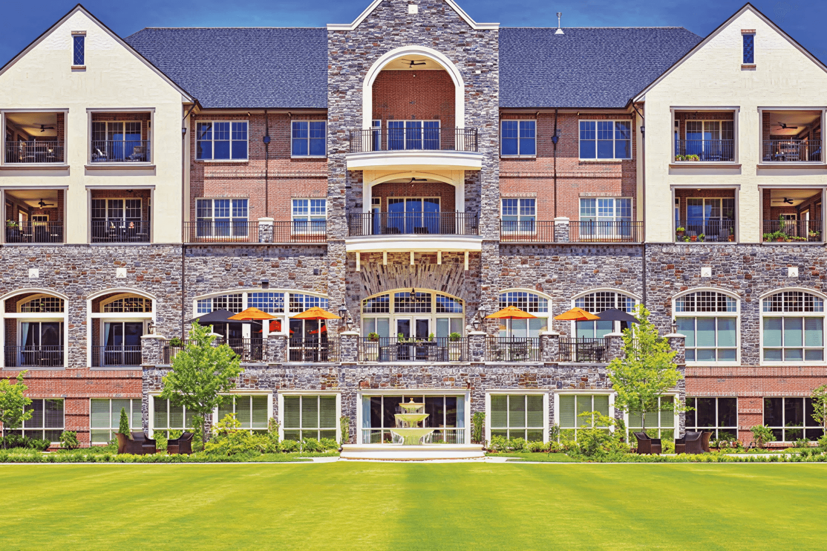 Multi-Story Residential Building Back View with Lawn Rear facade of a multi-story apartment building featuring stone and brick exterior, arched windows, balconies with orange umbrellas, and green lawn.