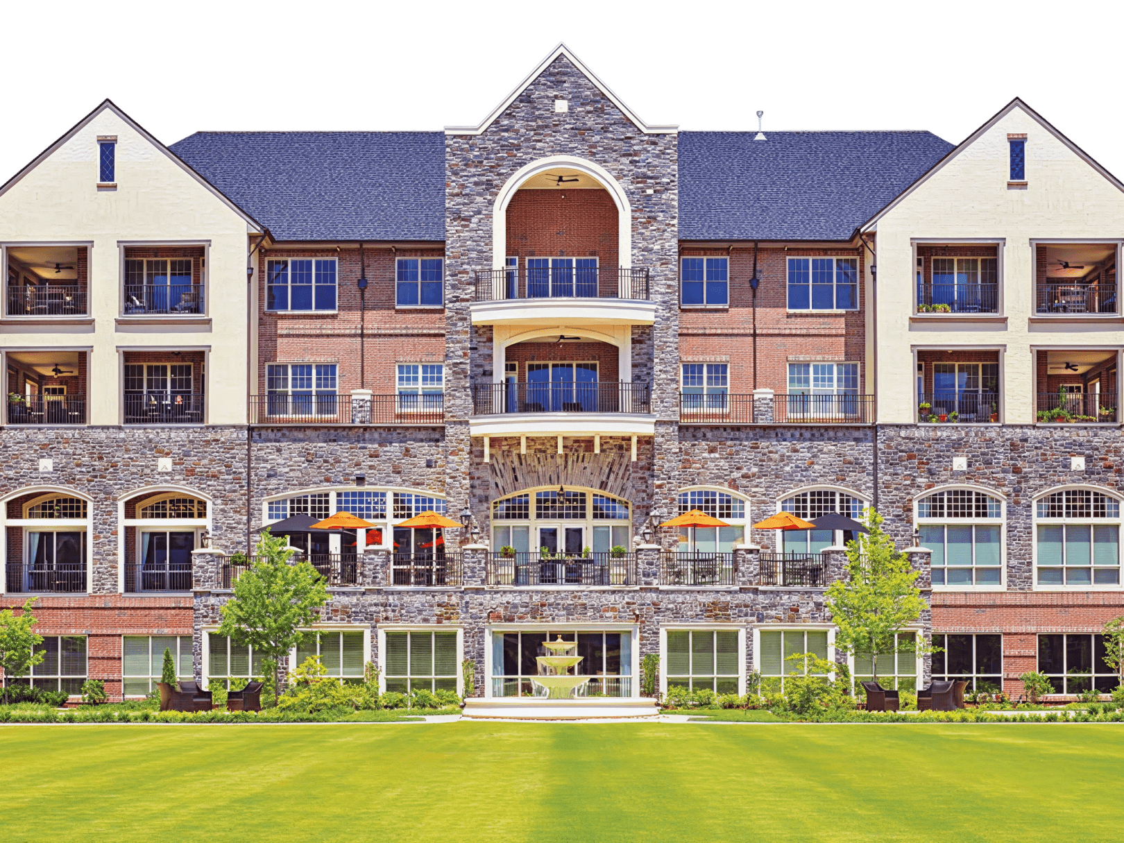 Front View of Multi-Story Mixed-Use Building Symmetrical multi-story building with stone and brick facade, featuring residential units, ground-floor commercial spaces with outdoor seating, and manicured lawn.