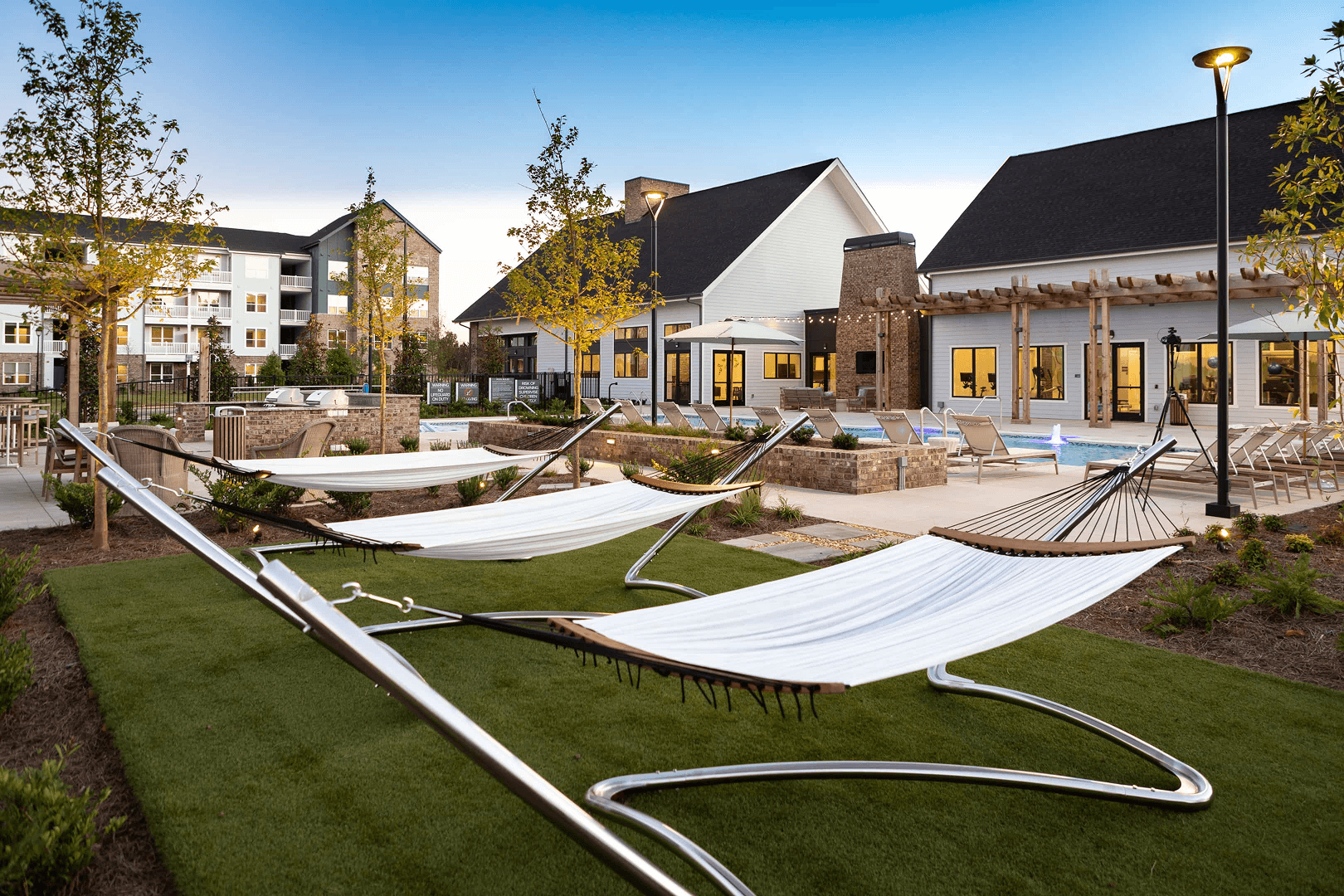 Luxury Apartment Complex Pool and Hammock Area Modern apartment complex outdoor amenities featuring white hammocks on lawn, swimming pool, pergola, and clubhouse with warm lighting at dusk