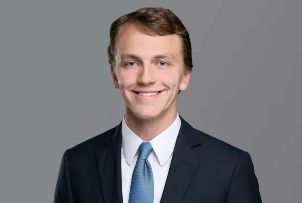 Professional headshot of Max Armstrong, a young man in a navy suit with blue tie, smiling at camera against gray background