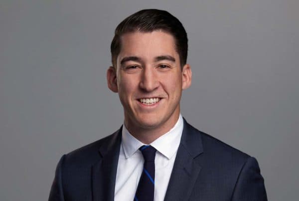 Professional headshot of Baker J smiling in a navy suit with white shirt and blue striped tie against gray background