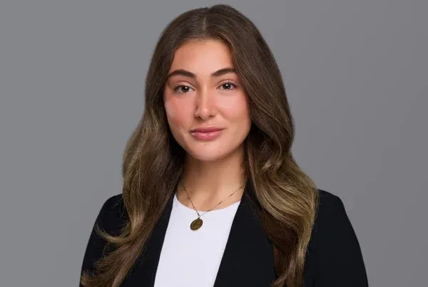 Professional headshot of Billi Brandner wearing a black blazer over white top with a necklace against gray background