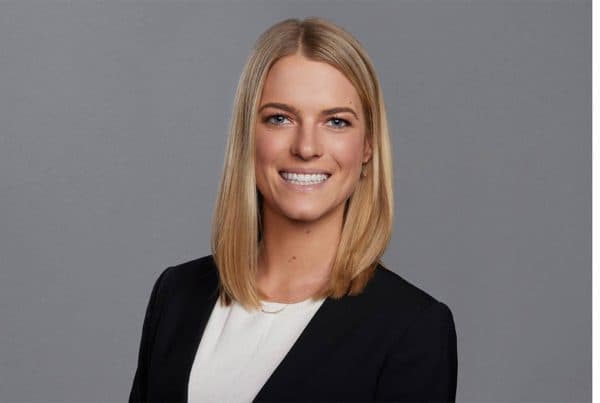 Professional headshot of a smiling blonde woman in a black blazer and white shirt against a gray background.