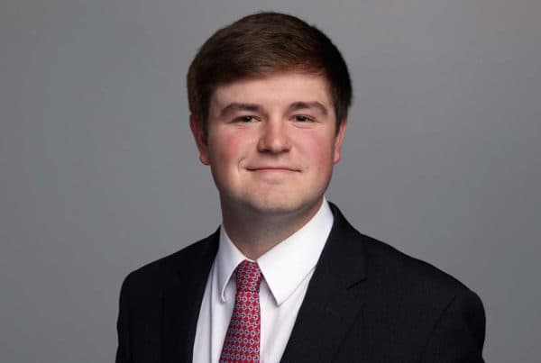 Professional headshot of Will Calhoun wearing a dark suit, white shirt, and red patterned tie against a gray background.