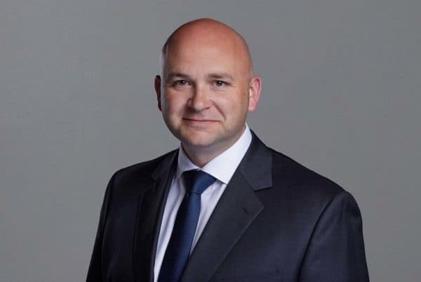 Professional headshot of a bald businessman in a dark suit and blue tie against a gray background