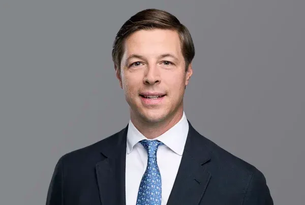 Professional headshot of Will Corbitt wearing a navy suit, white shirt, and blue patterned tie against a gray background