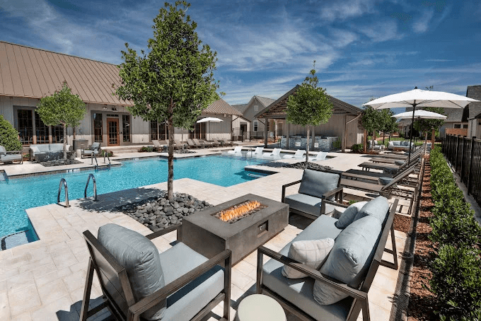 Modern resort-style pool with lounge chairs, fire pit seating area, and residential buildings under blue sky