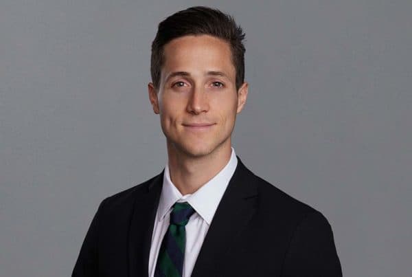 Professional headshot of Drake C, a young man in a black suit with white shirt and dark green tie, smiling at camera against gray background.