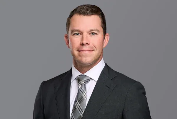 Professional headshot of Adam Duerr in dark suit and plaid tie against gray background
