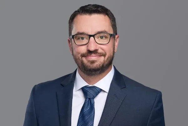 Professional headshot of Shane Grohs wearing glasses, beard, navy suit and striped tie against gray background
