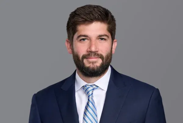 Professional headshot of James Hanna wearing a navy blue suit, white shirt, and blue striped tie against a gray background.