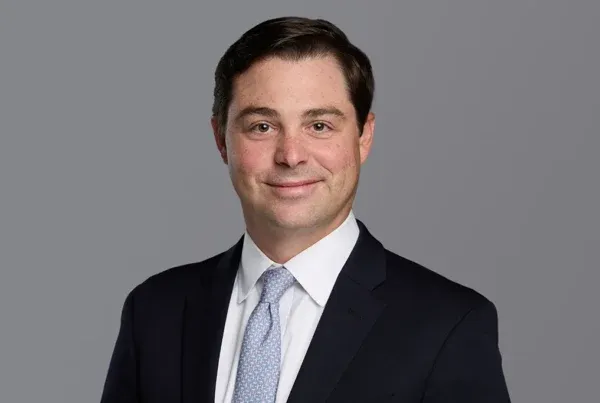 Professional headshot of Jake Harris wearing a dark suit, white shirt, and light blue patterned tie against a gray background.