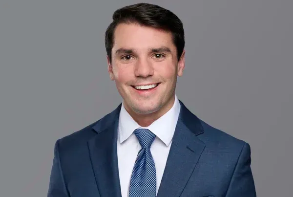 Professional headshot of Heath Cooper wearing a navy blue suit, white shirt, and patterned blue tie, smiling at camera.