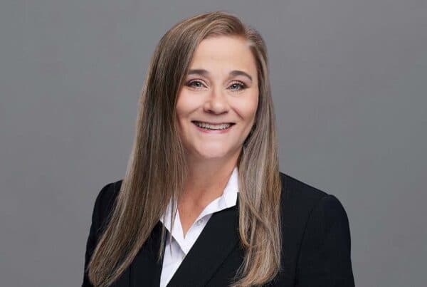 Professional headshot of Holland R. smiling, wearing a black blazer and white collared shirt against a gray background.