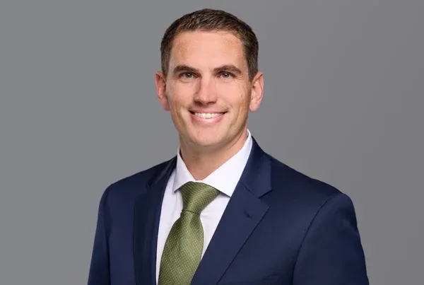 Professional headshot of Brad Kaaber wearing a navy suit, white shirt, and olive green tie against a gray background.