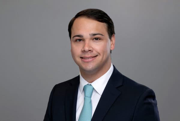 Professional headshot of King Will wearing a navy suit, white shirt, and light blue tie against a gray background