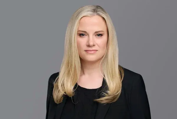 Professional headshot of Kristin Koenig with blonde hair wearing a black blazer against a gray background
