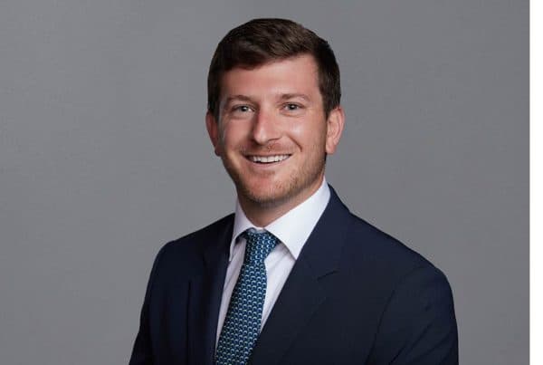Professional business portrait of a smiling man in a navy suit and blue patterned tie against a gray background.