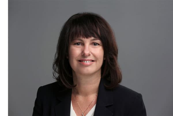 Professional headshot of Alexandra Logan, a woman with shoulder-length dark hair wearing a black blazer, smiling at camera against gray background.