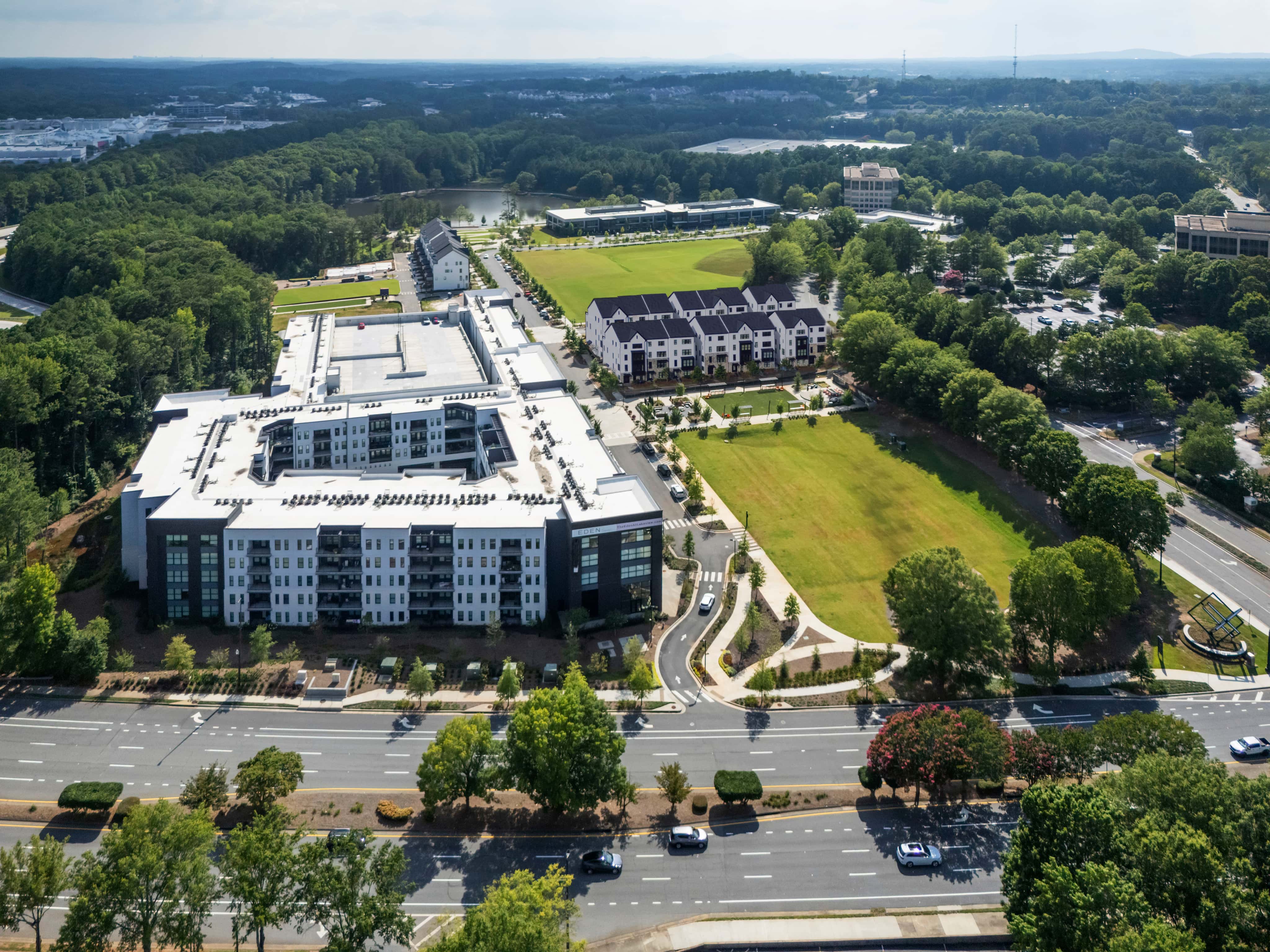 Aerial photograph showing modern apartment buildings, green spaces, curved roads, and landscaping in a master-planned residential community surrounded by trees.