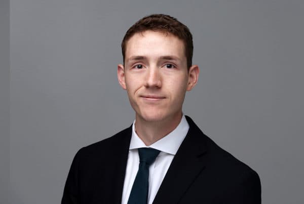 Professional headshot of Kennan O'Brien wearing a black suit, white shirt, and dark tie against a gray background.