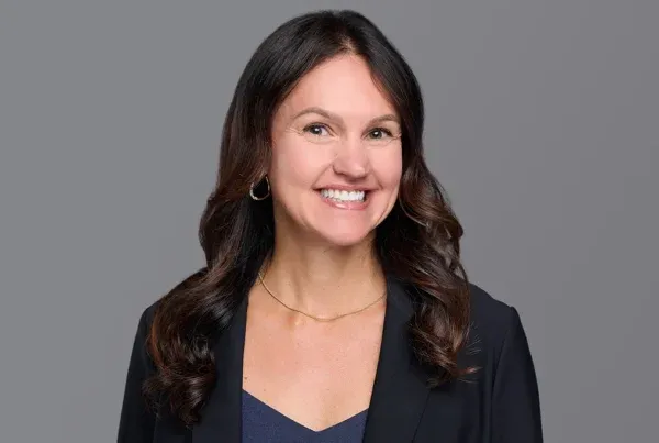 Professional headshot of Melissa Perignat smiling, wearing a black blazer and navy top against a gray background.
