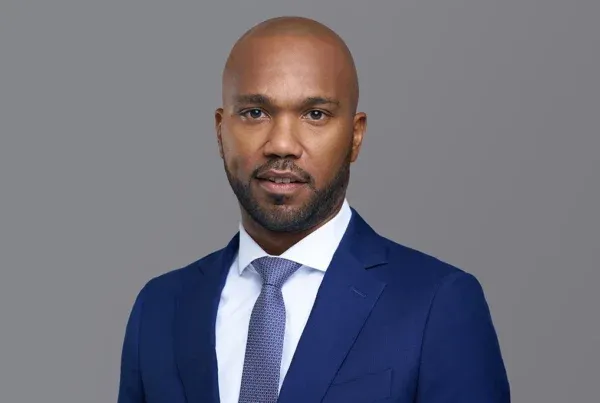 Professional headshot of Rashid A., a bald man with beard wearing a navy blue suit and patterned tie against gray background