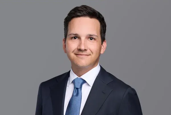 Professional headshot of Dominic Reinecker in a dark navy suit with blue tie against a gray background