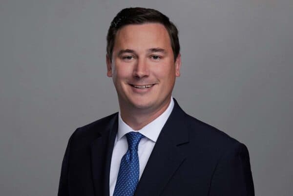 Professional headshot of a smiling man in a dark suit with blue tie against a gray background
