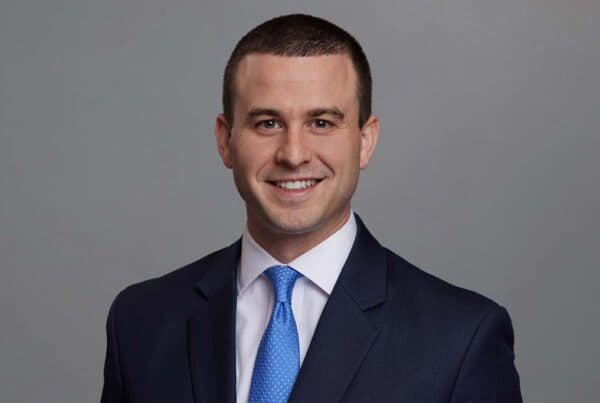 Professional headshot of Steven Rudel wearing a dark suit, light blue shirt, and blue tie against a gray background