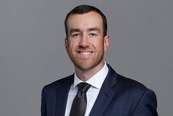 Professional business portrait of a smiling man in a navy suit and tie against a gray background