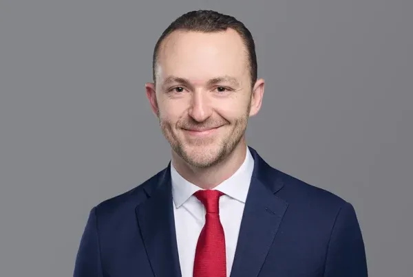 Professional headshot of Andrew Sorensen wearing a navy suit, white shirt, and red tie against a gray background