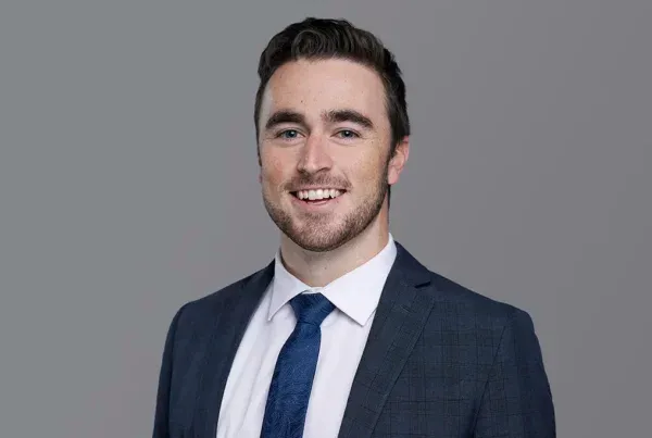 Professional headshot of a smiling man named Nick wearing a navy suit, white shirt, and blue tie against a gray background.