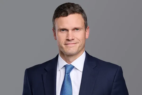 Professional headshot of Tyler Wakefield wearing a navy suit, white shirt, and blue patterned tie against a gray background