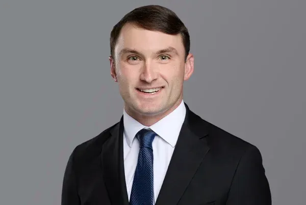 Professional headshot of Isaac Weeks wearing a dark suit, white shirt, and blue tie against a gray background.