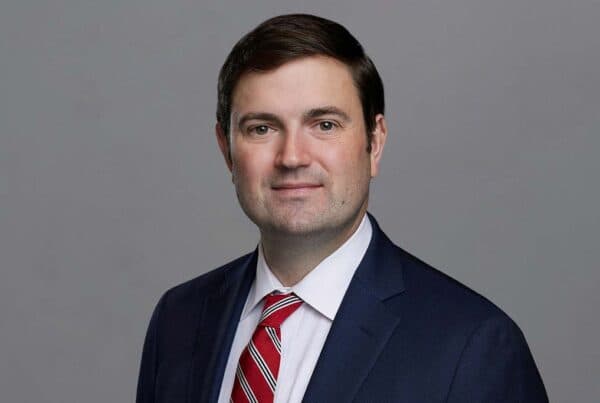 Professional headshot of Weston W. wearing a navy suit, white shirt, and red striped tie against a gray background.