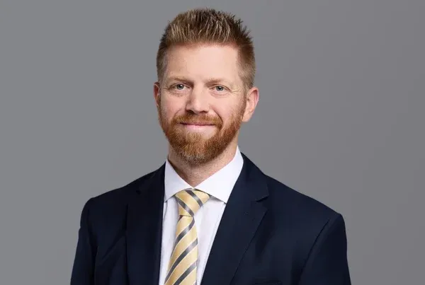 Professional headshot of Elliot Williams, a man with a red beard wearing a navy suit and striped tie against a gray background