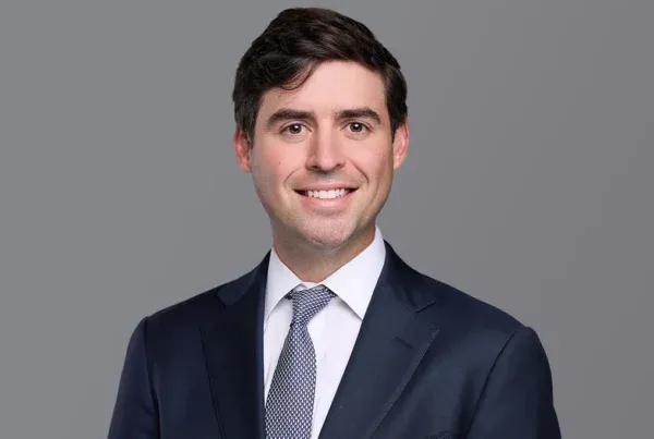 Professional headshot of Luke Zemis wearing a navy suit, light blue shirt, and patterned tie against a gray background
